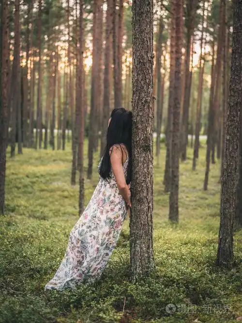 夕阳下的绿色森林中的孤独女子穿着站立和倚在树上