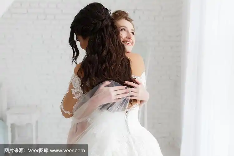 奢华的新娘拥抱着伴娘微笑,在极简的阁楼白砖背景下的快乐时刻