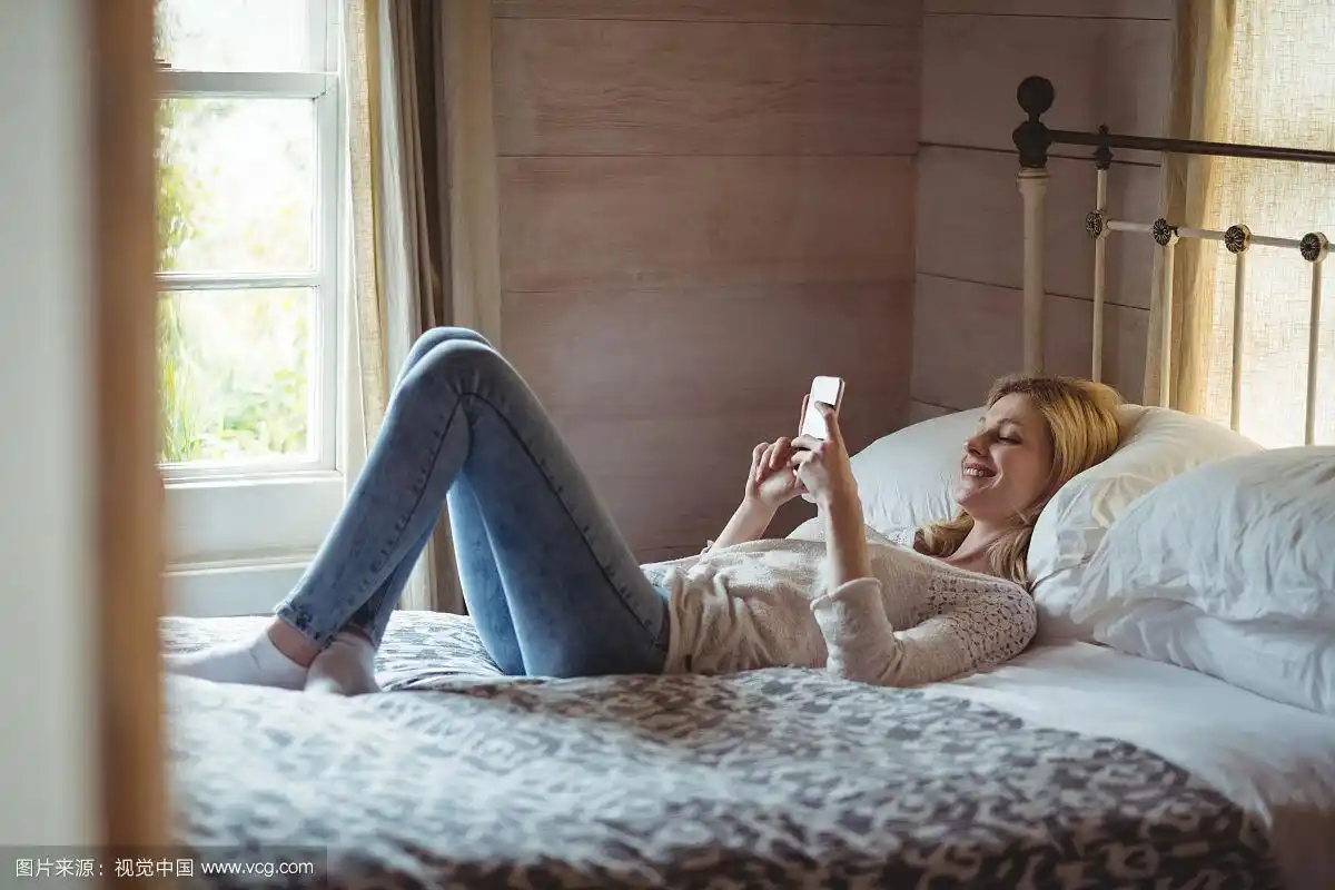 美丽的女人躺在床上,在卧室里使用手机