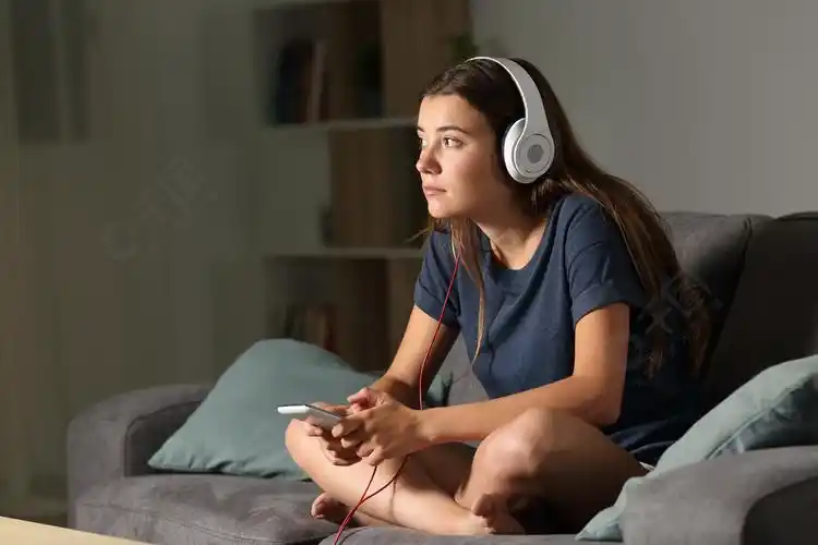 一个沉思的青少年在晚上坐在家里客厅的沙发上听音乐的全身肖像