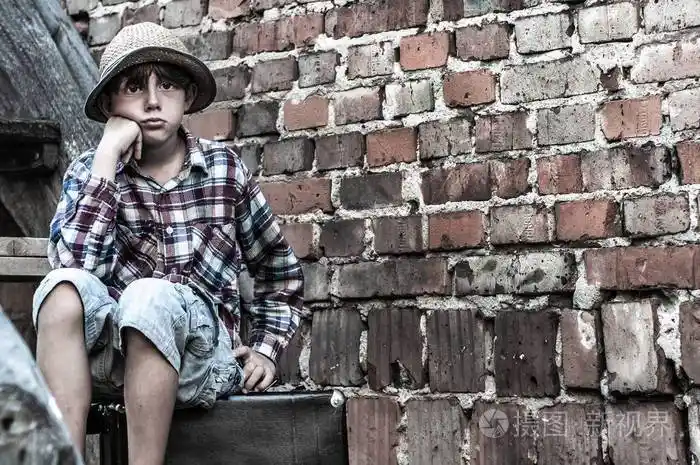 可怜的男孩靠着砖墙坐在手提箱上无家可归的孤独的孩子