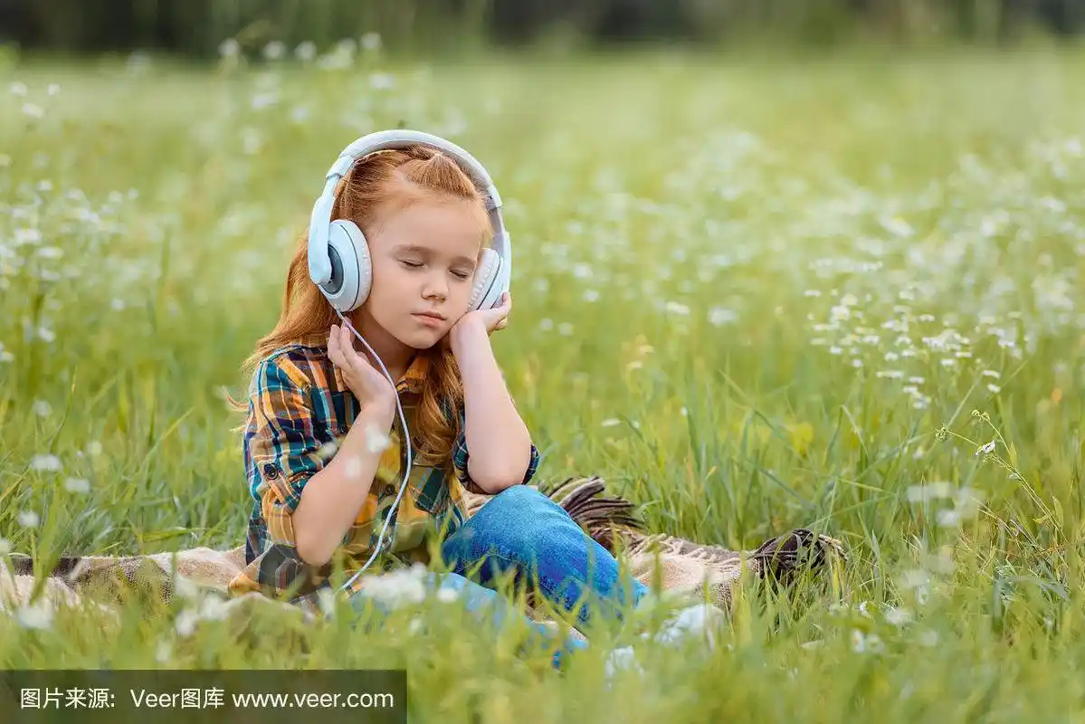 躺在草地上的毯子上,闭着眼睛戴着耳机听音乐的小孩