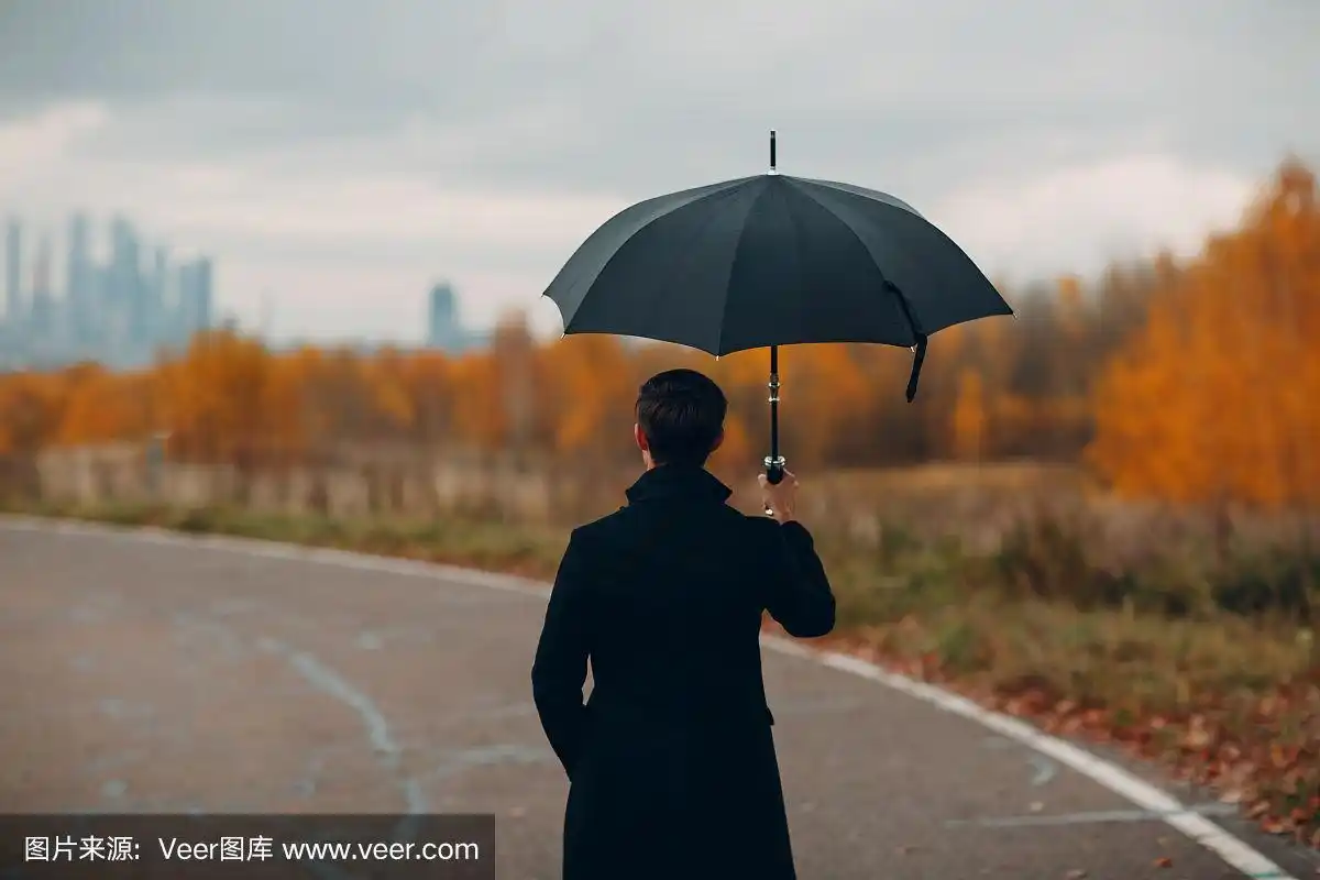 从背影看,一个穿着黑色外套的年轻人打着伞在雨中走向城市