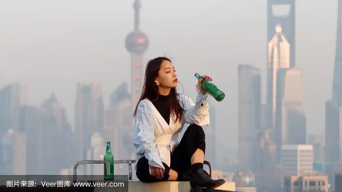 美丽的年轻女子坐在大厦屋顶上,手里拿着一个空啤酒瓶,背景是模糊的