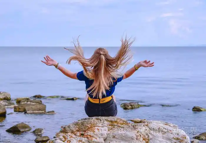 夹克的女孩她的头发朝下坐在平静的海面上的一块石头上回头望着远处