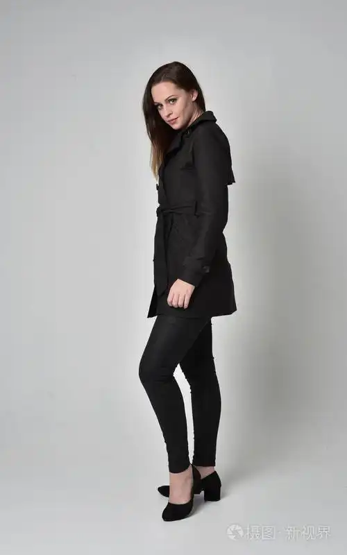 一个黑发女孩穿着黑色长外套,站在侧面轮廓灰色工作室背景的完整长度