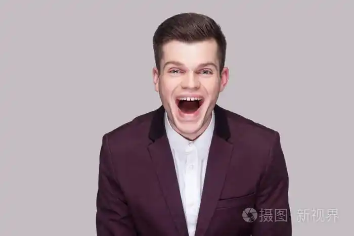 滑稽英俊的年轻人穿着紫罗兰色西装和白色衬衫,张开嘴看着相机,在灰色