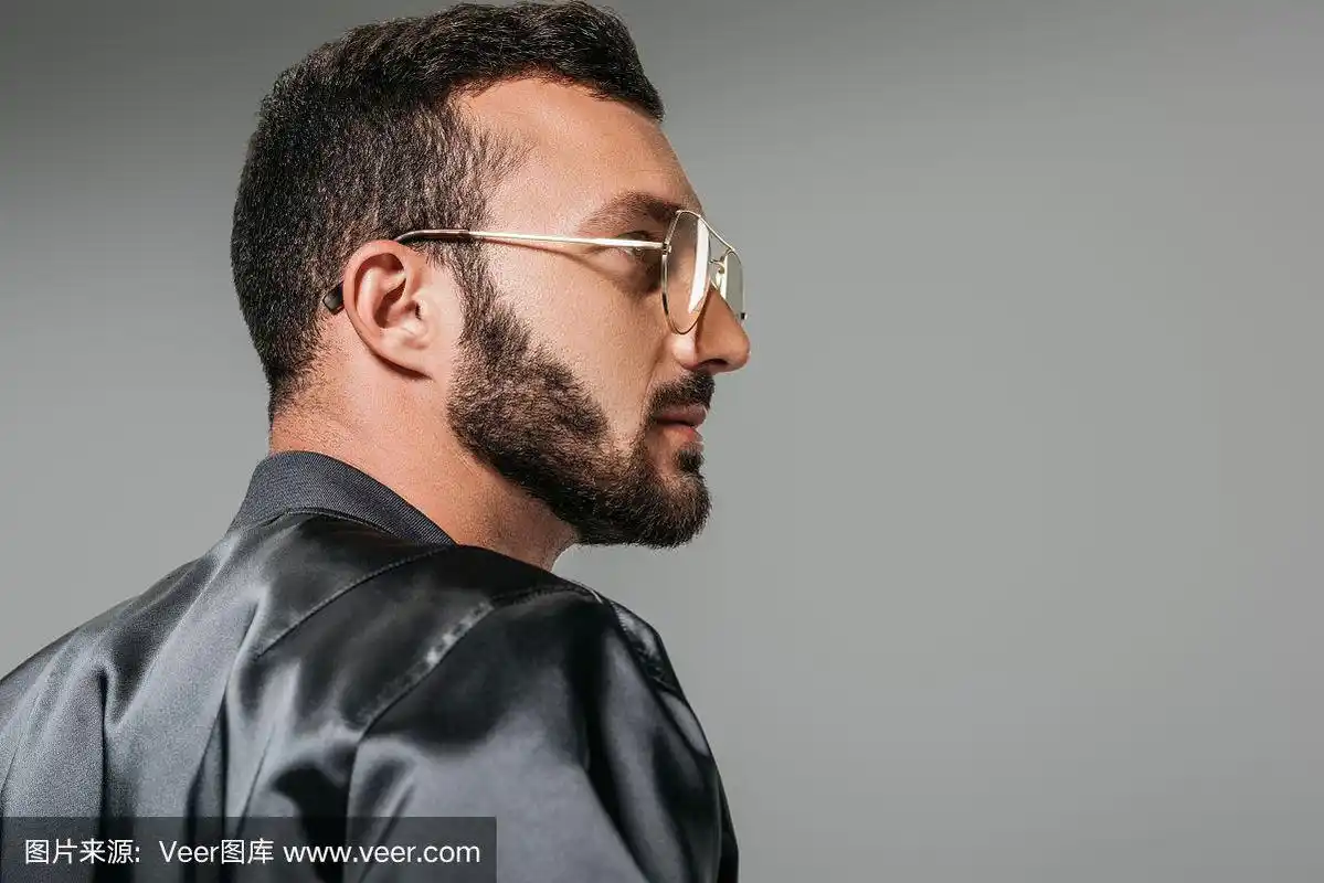 侧面的胡子男子在时尚的眼镜和黑色轰炸机孤立在灰色背景