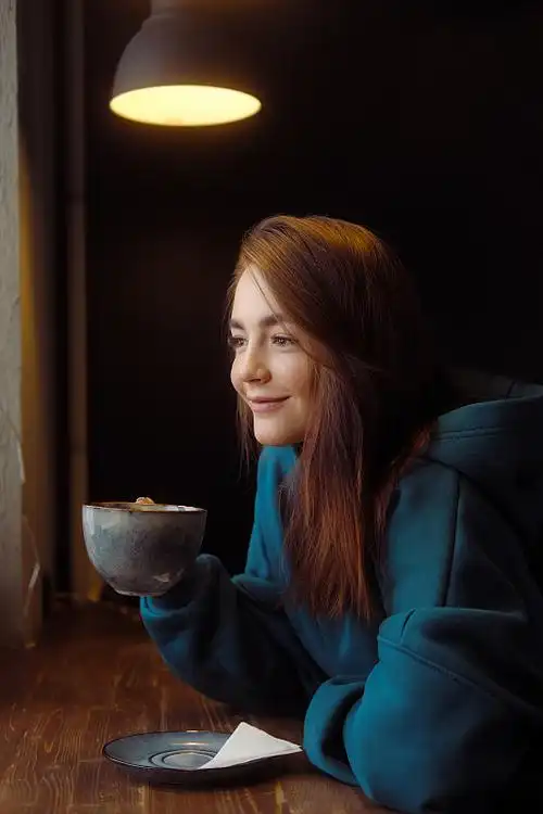 一个穿着蓝色连帽衫的漂亮女孩坐在咖啡桌旁.女人用一个大杯子喝咖啡.