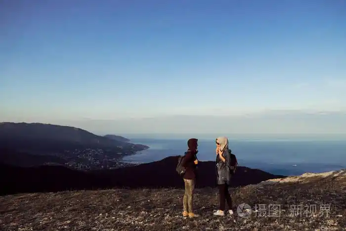 两个女孩旅行者背对着山顶边的镜头站着