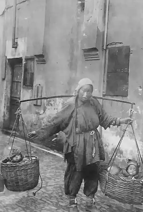 清末照片,一位母亲用扁担挑着一对箩筐,两个孩子戴着虎头帽,坐在筐中